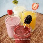 4 smoothies in tall glasses on a wicker table. Smoothie colours are purple, pink, white and yellow