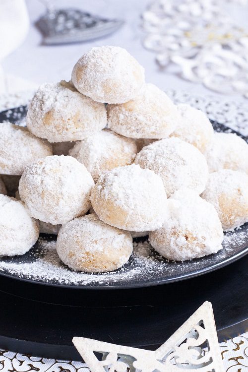 A stack of ball-shaped cookies on a black plate. It is dusted generously with powdered sugar so they are white as snow balls. White and silver christmas ornaments are placed next to them.